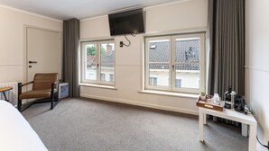 A hotel room with a bed, a chair, a small table with a tray, a TV mounted on the wall, and large windows with curtains. - Contemporary Room in Bruges' Heart (Brugge)