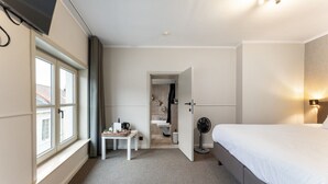 A hotel room with a bed, a small table with a plant, a fan, and a view of the outside through a window. - Contemporary Room in Bruges' Heart (Brugge)