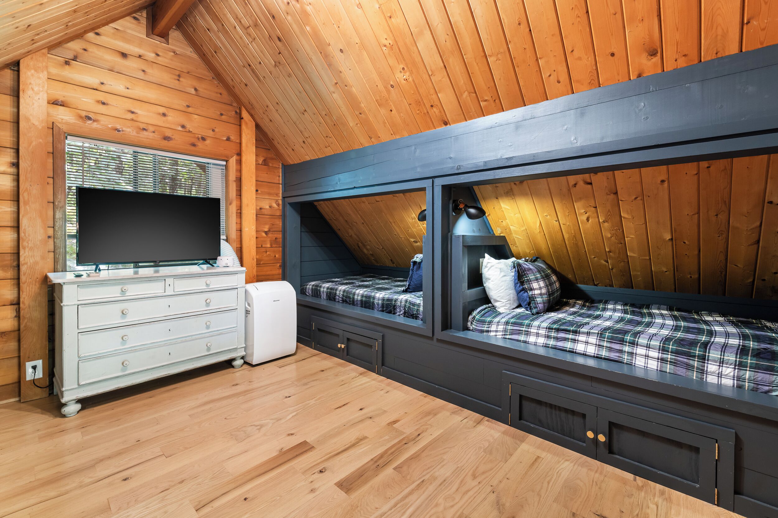 Ein Zimmer mit Holzwänden und geneigter Decke, mit einem Bett mit kariertem Überwurf, einer Kommode mit Schubladen und einem Flachbildfernseher auf einem Holzboden.