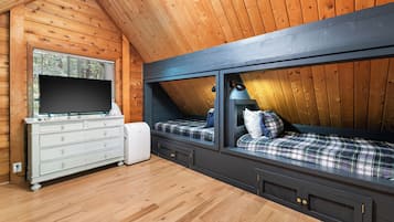 Ein Zimmer mit Holzwänden und geneigter Decke, mit einem Bett mit kariertem Überwurf, einer Kommode mit Schubladen und einem Flachbildfernseher auf einem Holzboden.