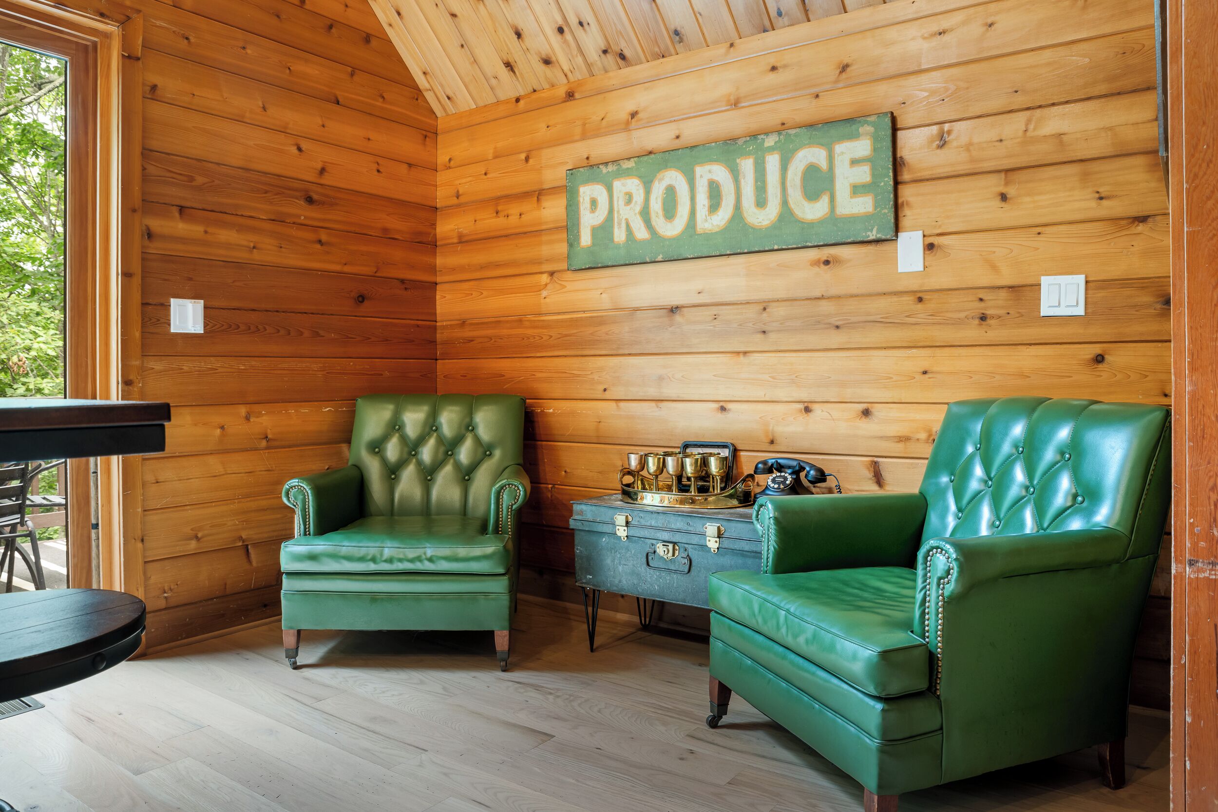 A cabin interior with two green leather armchairs, a wooden wall with a 'PRODUCE' sign, and a vintage suitcase.