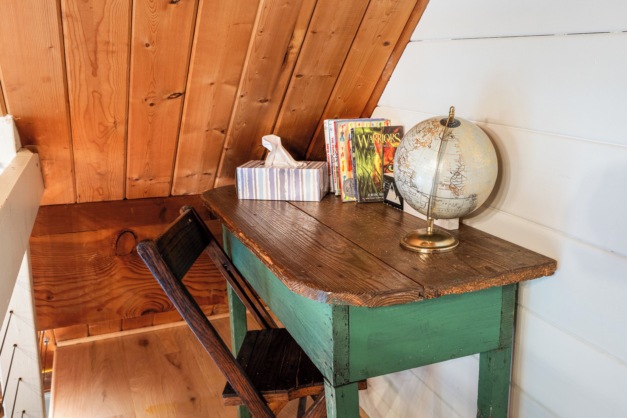 A wooden desk with a globe, books, and a tissue box in a room with wooden walls and a sloped ceiling.
