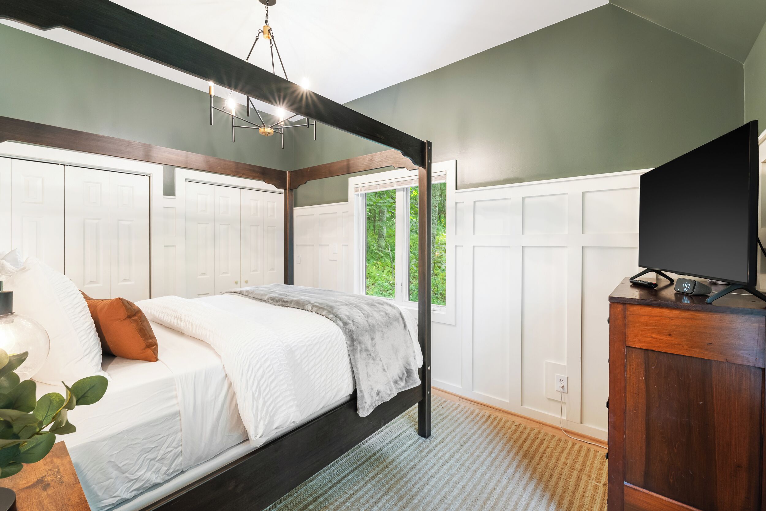 A bedroom with a four-poster bed, a television on a wooden cabinet, and a view of greenery through a window.