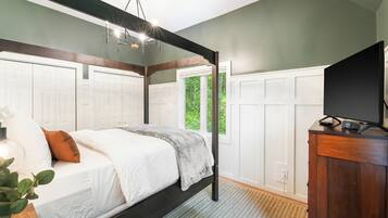 A bedroom with a four-poster bed, a television on a wooden cabinet, and a view of greenery through a window.