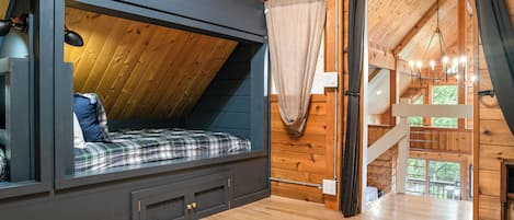 Un dormitorio tipo loft con techo inclinado, una cama con ropa de cama escocesa, un aparador oscuro y vistas al exterior a través de una ventana.