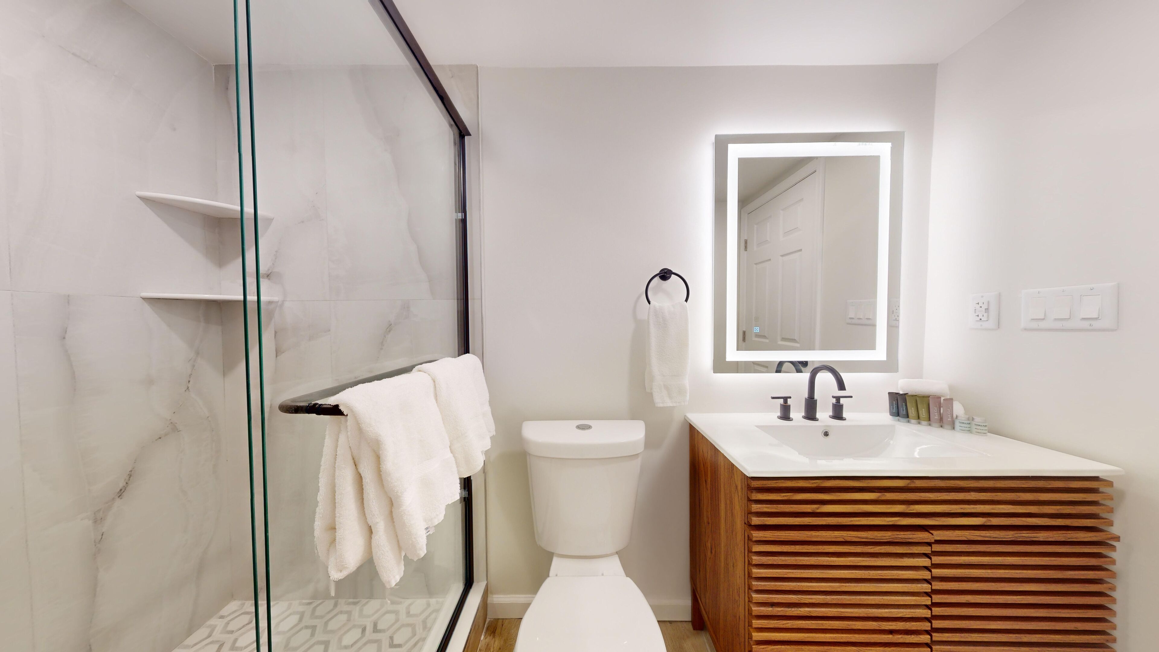 A modern bathroom with a glass shower enclosure, a white toilet, and a wooden vanity with a large mirror.