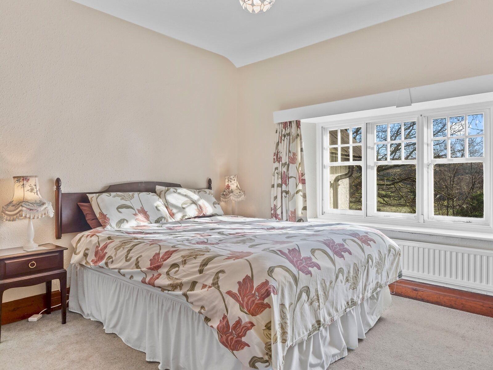 Een slaapkamer met een groot bed, nachtkastje, lamp en raam met uitzicht op bomen.