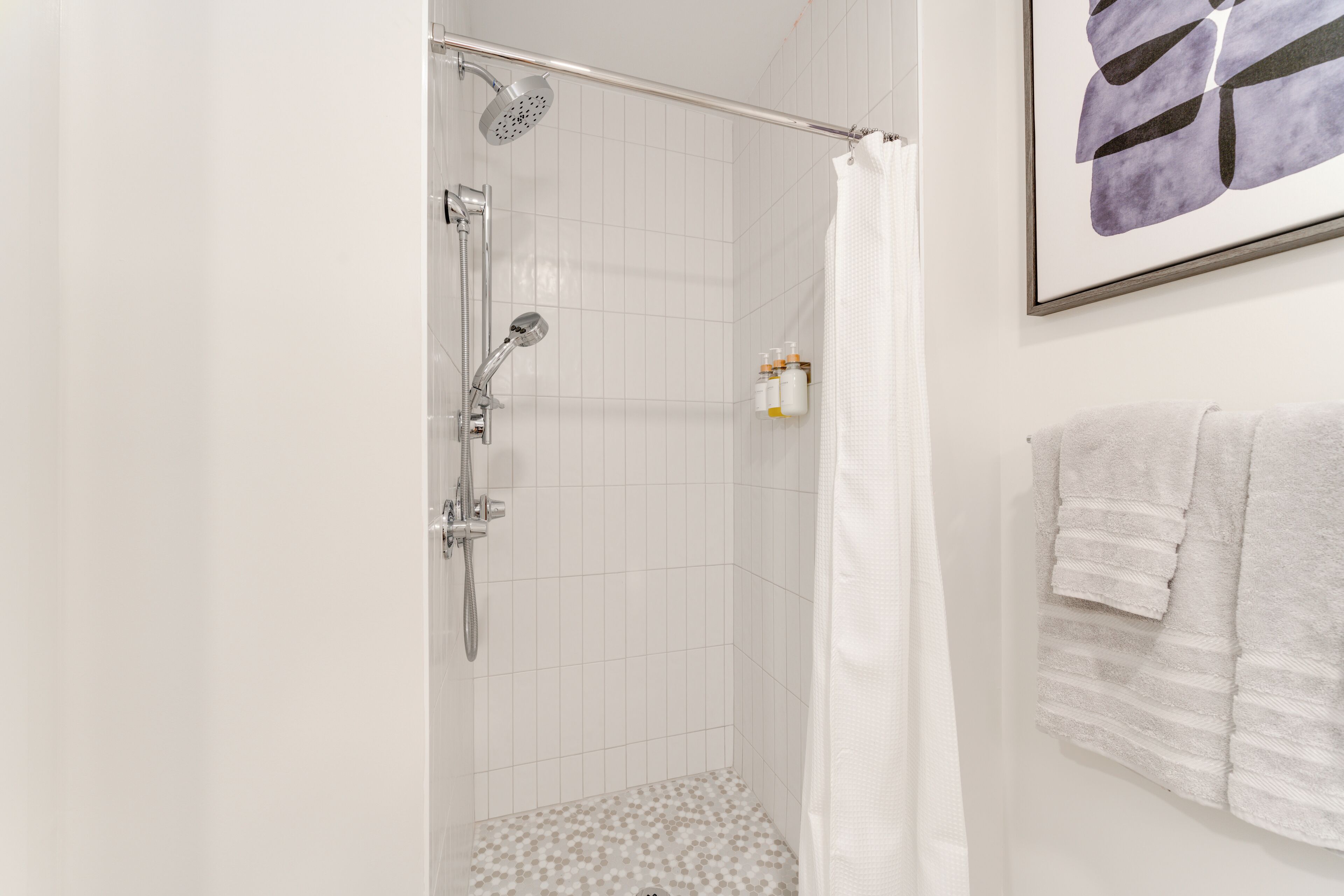 A shower with a handheld showerhead and a wall-mounted faucet, a tiled shower floor, and a framed abstract painting on the wall.