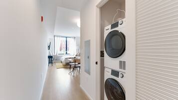 A laundry room with two stacked washing machines and a dryer, a wooden floor, and a view into a living area with a sofa and a dining table.