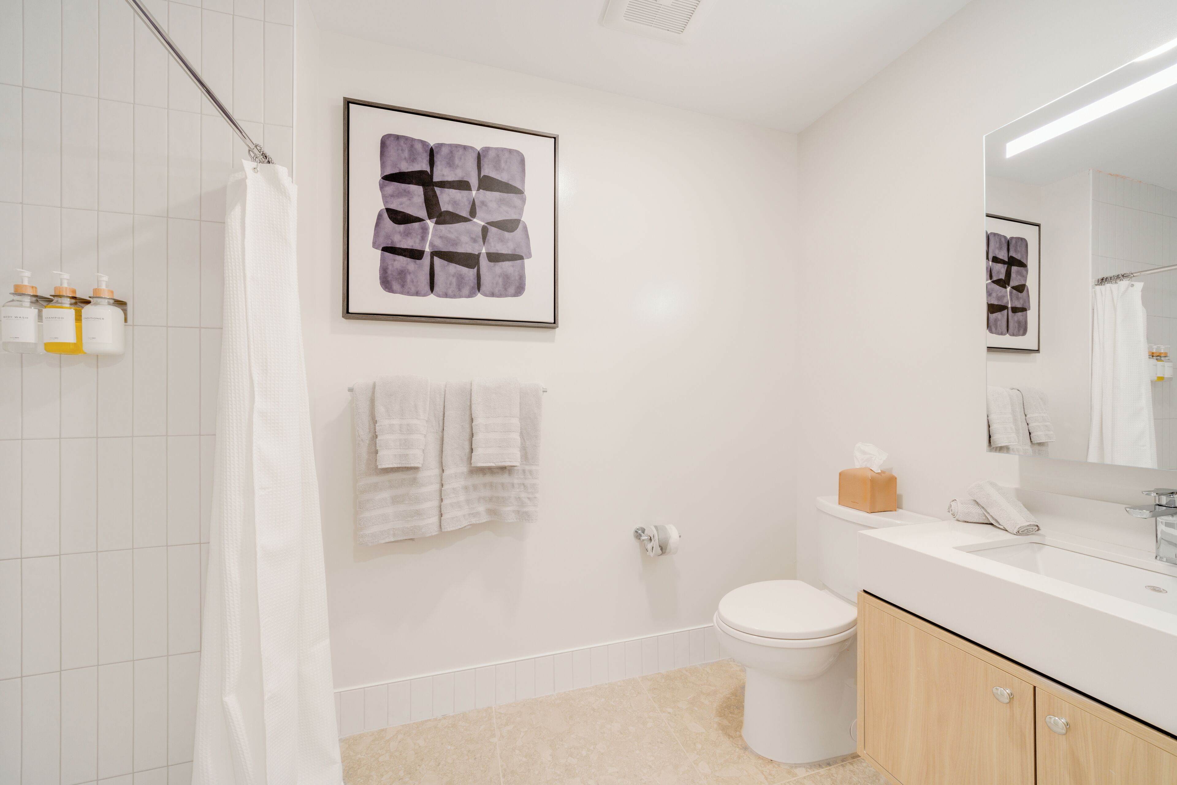 A modern bathroom with a white tiled wall, a beige floor, a white toilet, a wooden vanity with a mirror, and a framed abstract artwork above the mirror.