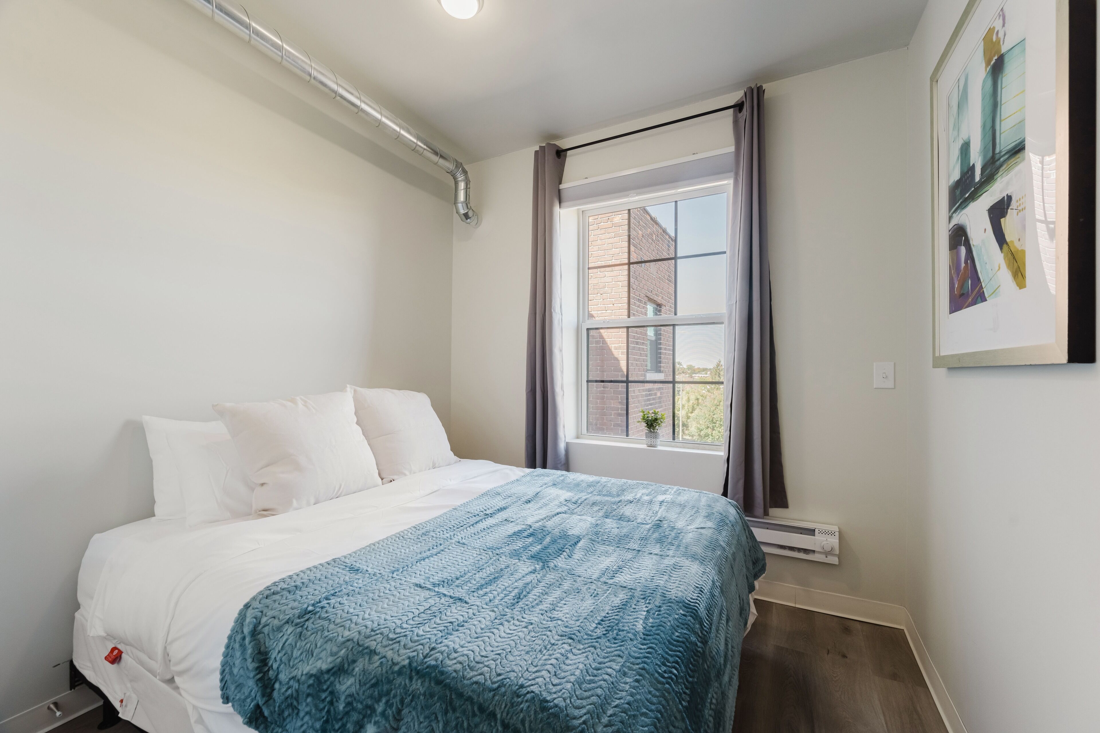 Een slaapkamer met een bed, een raam met gordijnen, een radiator en een aan de muur hangend schilderij.