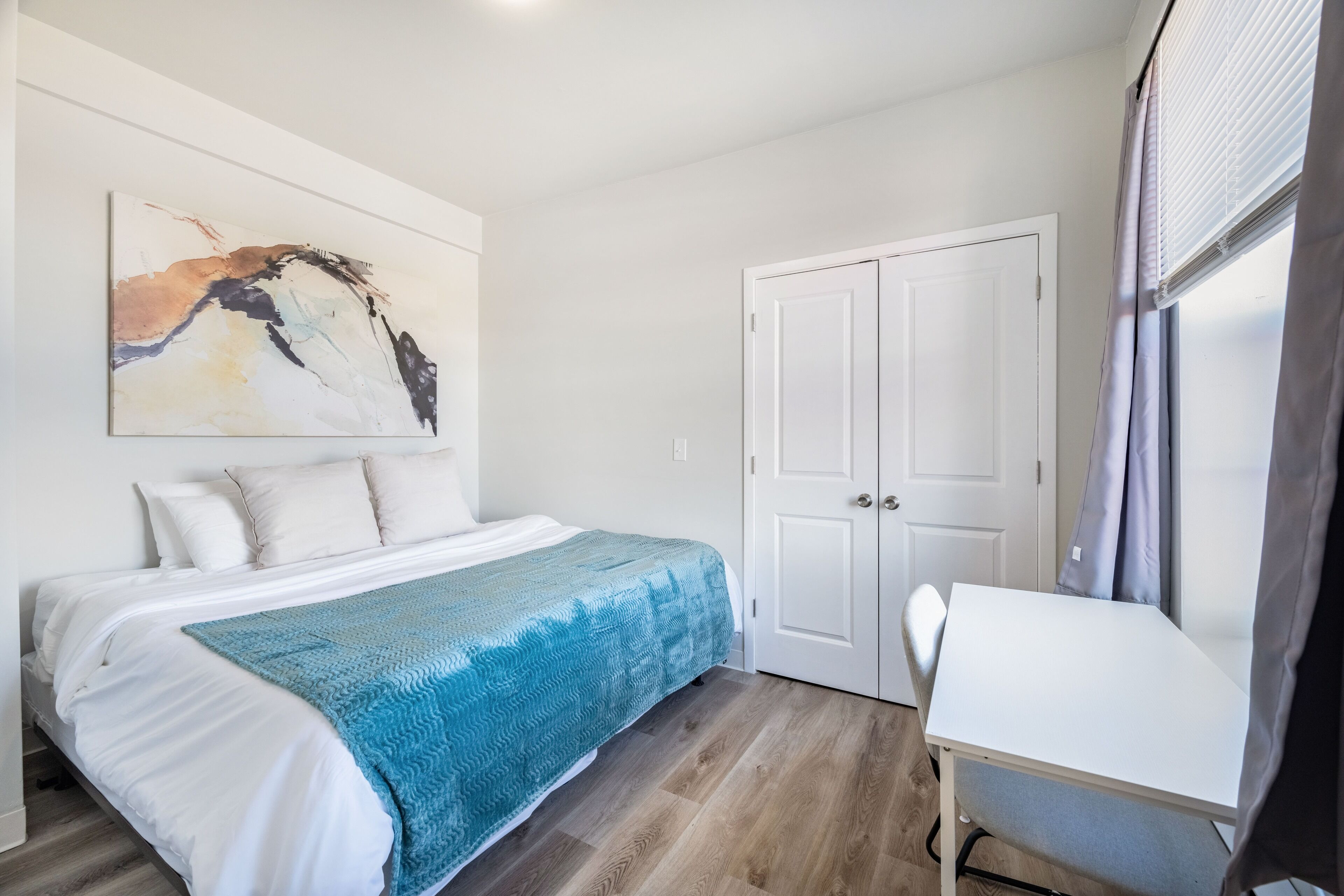 A bedroom with a bed, a desk, a door, and a wall with a painting.