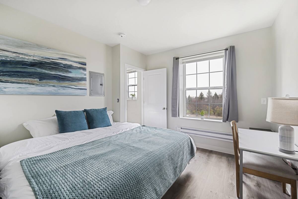 Une chambre à coucher avec un lit, un bureau, une chaise, une fenêtre avec des rideaux et un tableau accroché au mur.