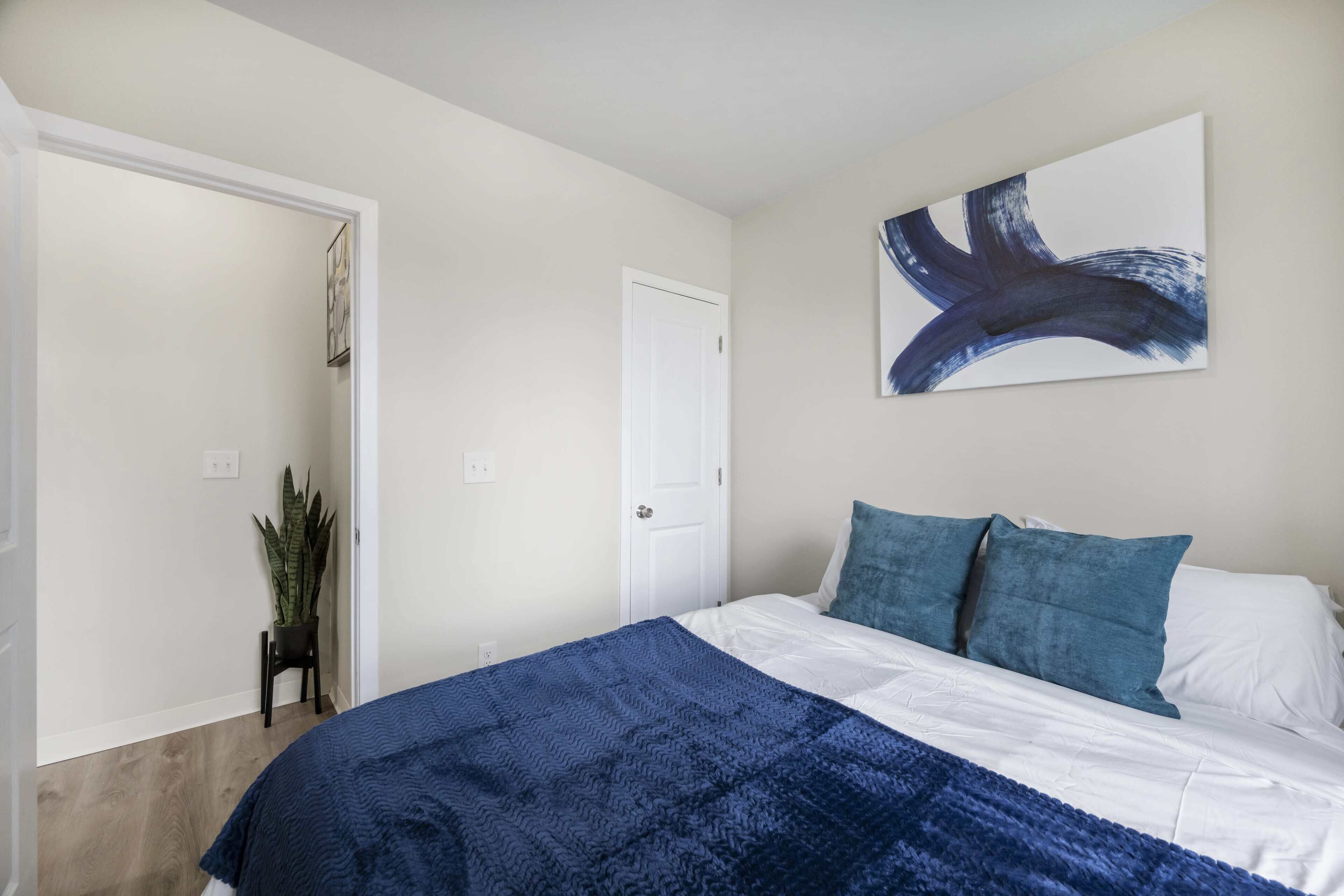 A bedroom with a bed, a door, a wall-mounted abstract painting, and a potted plant.