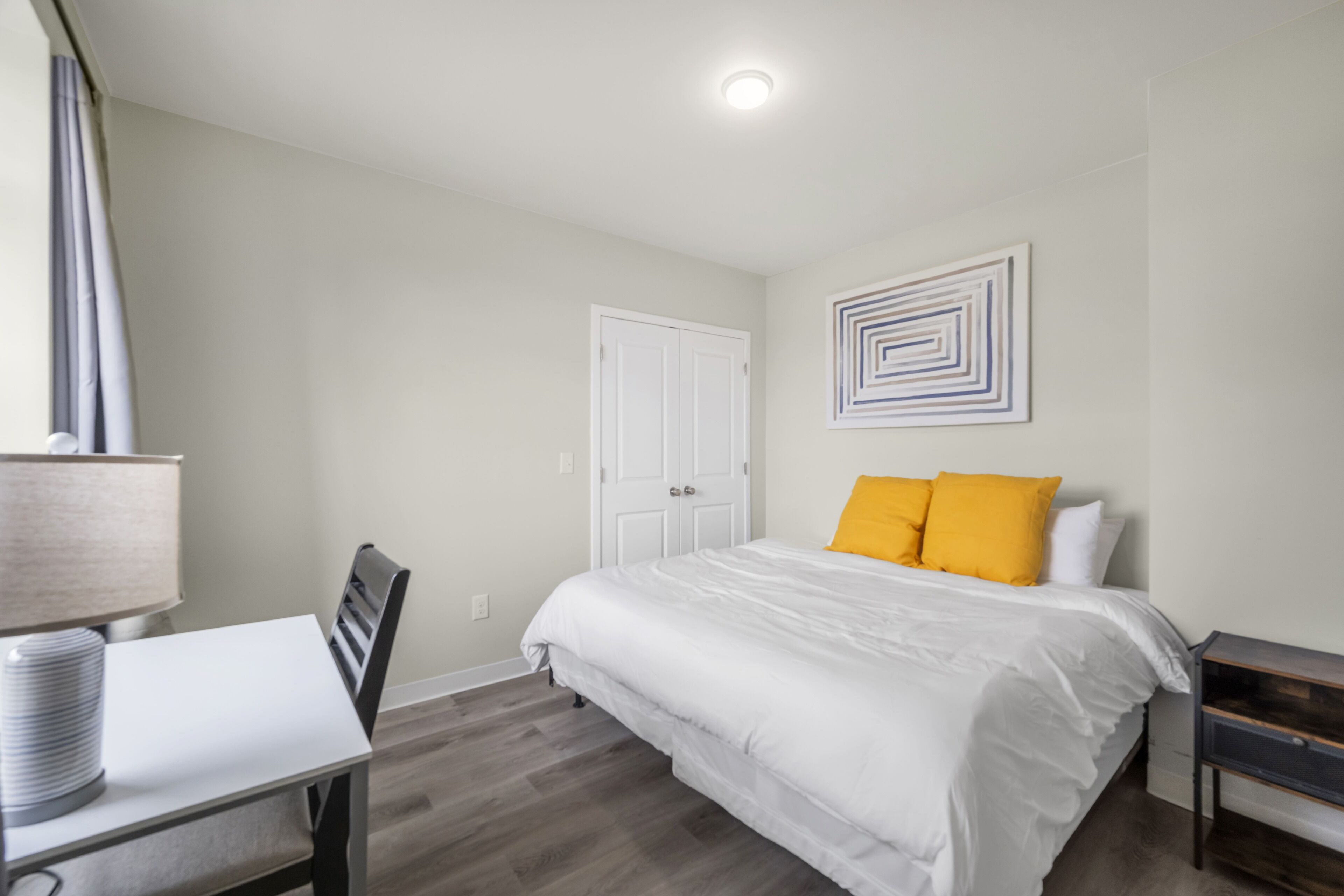 A bedroom with a bed, a desk with a lamp, a chair, a door, and a wall with a geometric art piece.