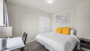 A bedroom with a bed, a desk with a lamp, a chair, a door, and a wall with a geometric art piece.
