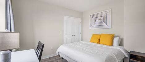 A bedroom with a bed, a desk with a lamp, a chair, a door, and a wall with a geometric art piece.
