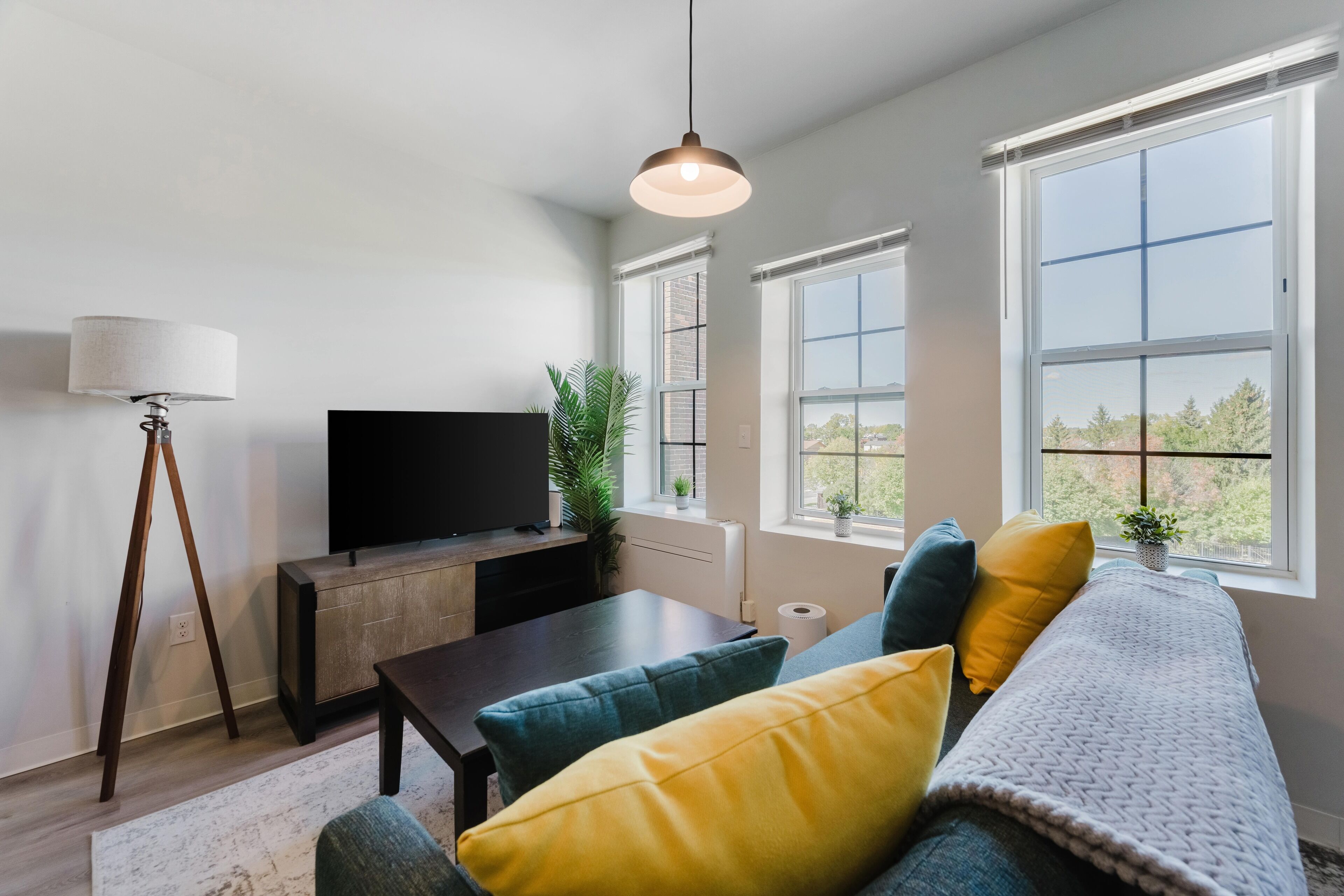 Ein modernes Wohnzimmer mit einem Flachbildfernseher, einem Couchtisch, einer Couch mit bunten Kissen, einer Stehlampe und großen Fenstern mit Blick ins Freie.