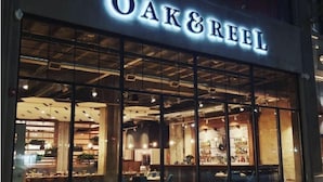 El exterior de un restaurante llamado “Oak & Reel”, con grandes ventanales de vidrio y un interior moderno visible a través de ellos.