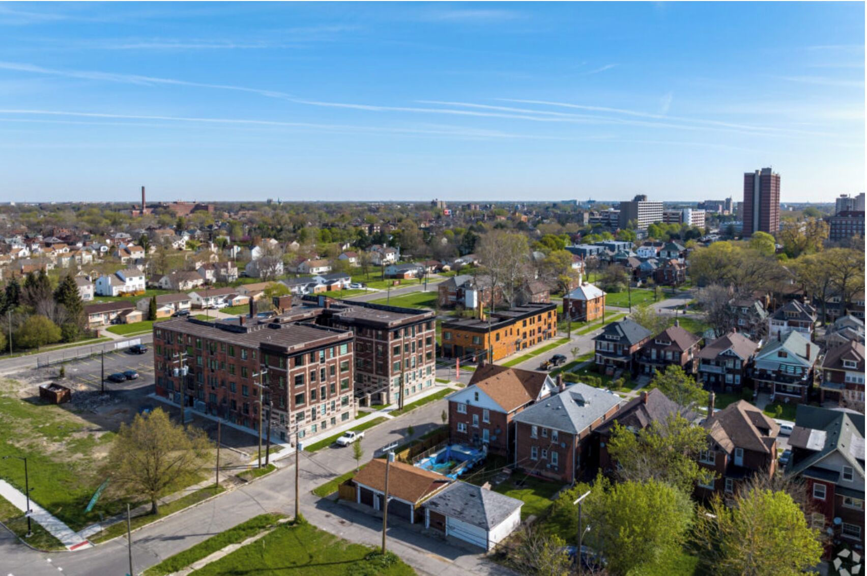 A residential area with a mix of single-family homes and multi-story buildings, a clear sky, and a distant view of a city skyline.
