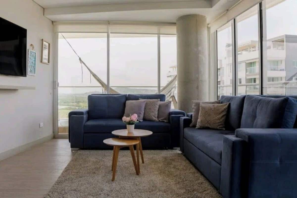 A modern living room with a dark blue sofa, a small round wooden table with a vase of flowers, and large windows offering a view of a bridge and buildings.