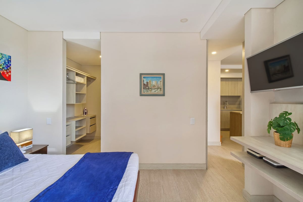 A bedroom with a bed, a TV, a painting, and a view into the kitchen.