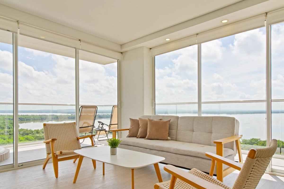 A spacious balcony with a sofa, chairs, and a table, overlooking a river and greenery.
