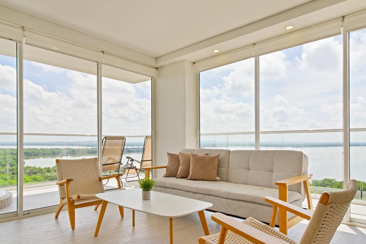 Ein geräumiger Balkon mit einer Couch, Stühlen und einem Tisch, mit Blick auf ein Gewässer und viel Grün.