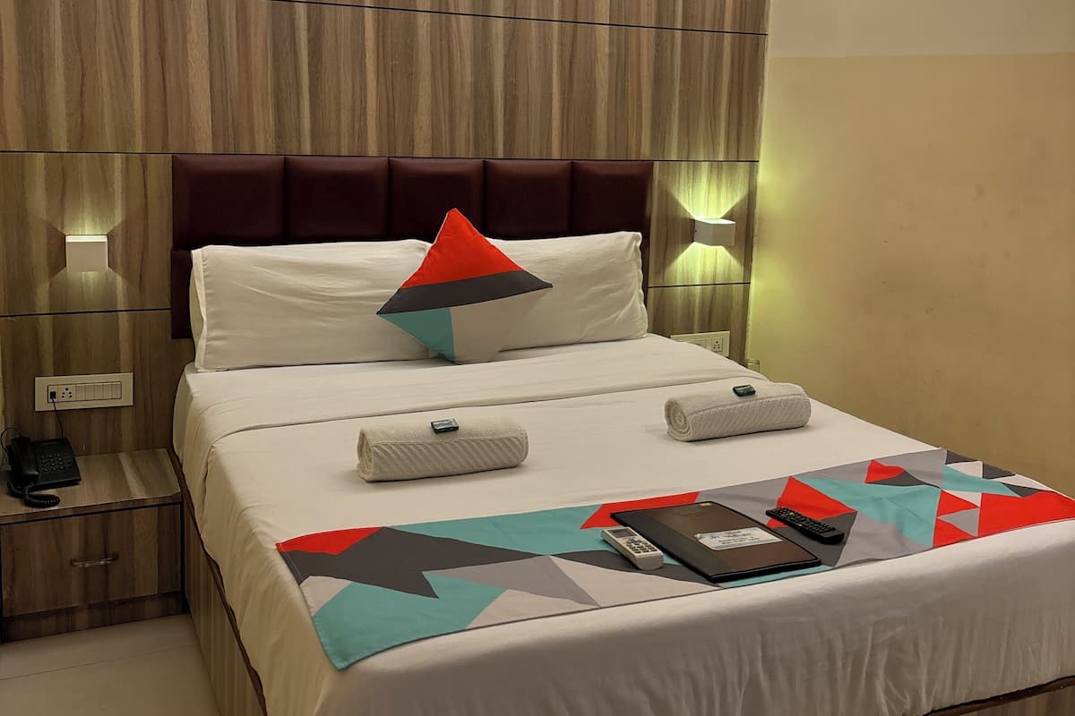Ein Hotelzimmer mit Holz-Kopfstütze, einem Bett mit weißer Bettwäsche, einem rot-schwarzen Kopfkissen, zwei gerollten Handtüchern, einem Nachttisch mit Telefon und einem Deckenventilator.