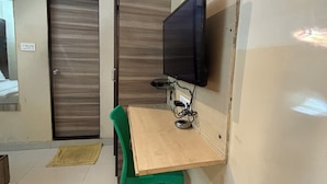 Una stanza piccola con una scrivania in legno, una sedia verde, una televisione a parete e una porta chiusa.