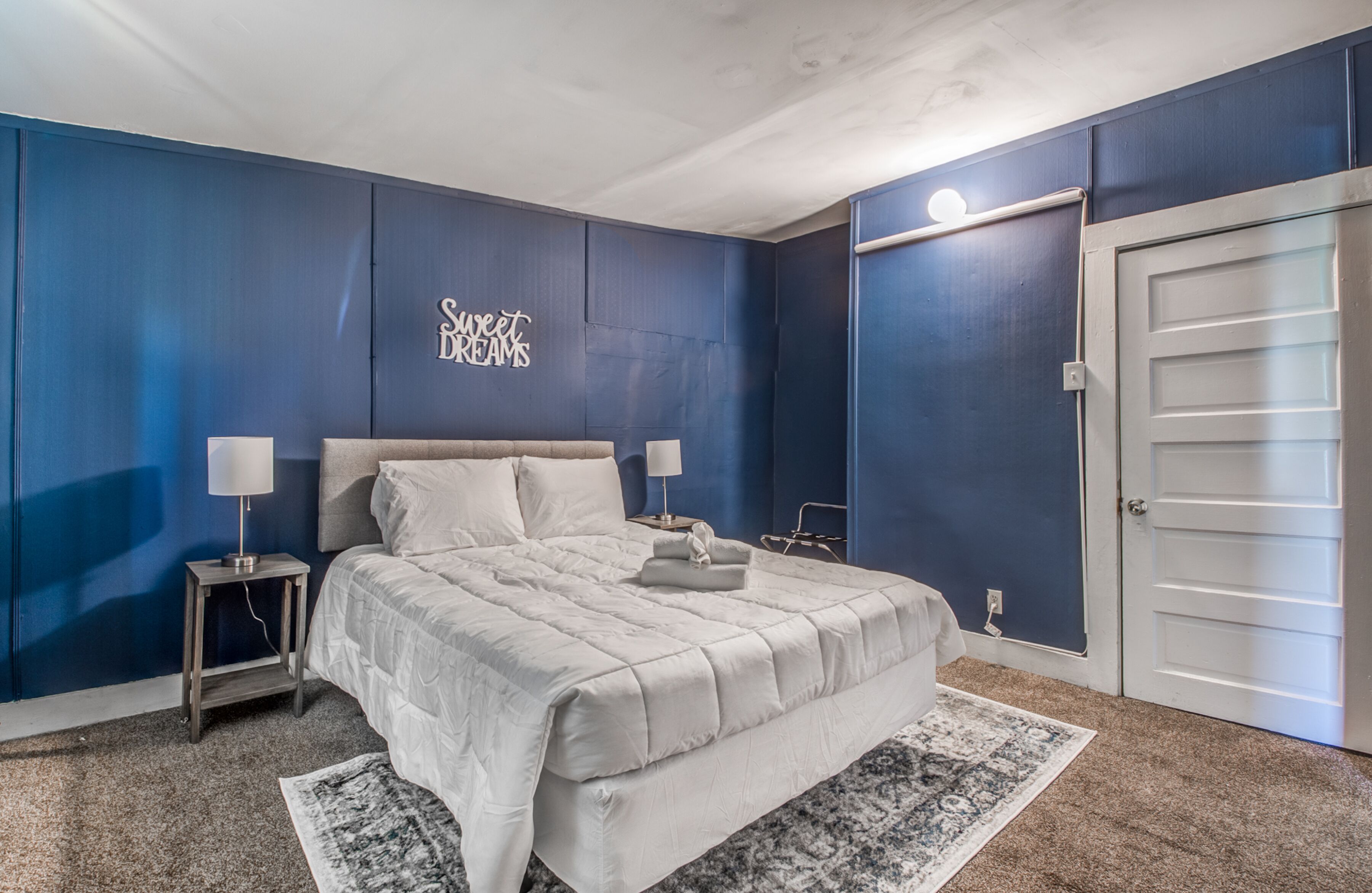 Un dormitorio con una cama grande, dos mesitas de noche con lámparas, un cartel en la pared y una puerta.