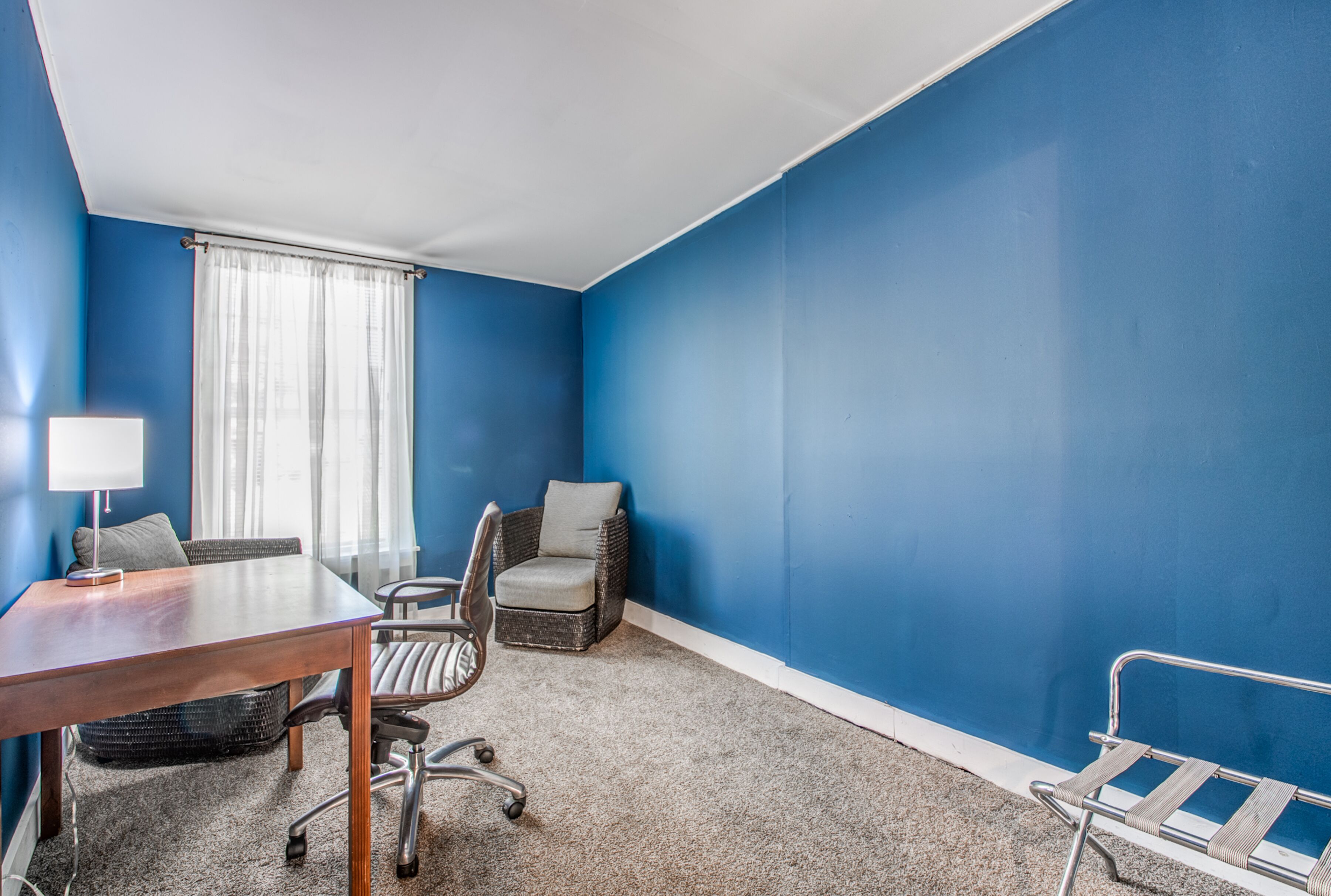 A room with blue walls, a wooden table, a chair, a small sofa, and a window with curtains.