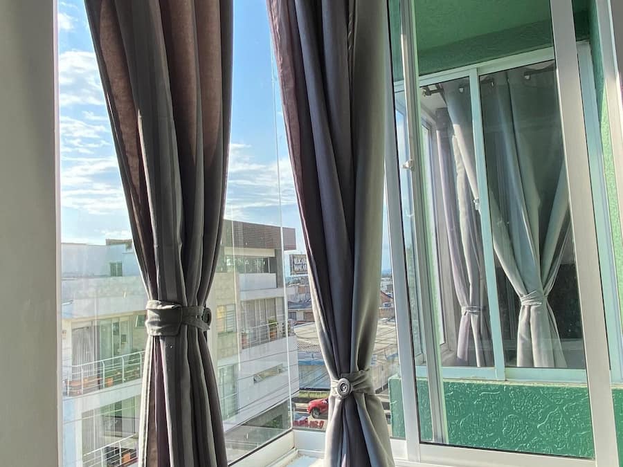 Quarto com janela e cortinas cinzentas amarradas, mostrando vista panorâmica da cidade.