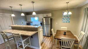A modern kitchen with a central island, white chairs, and a dining table. - Havens by the Sea STE A Cozy 2BR Beachside Escape (North Myrtle Beach)