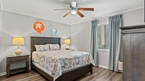 A bedroom with a bed, two bedside tables with lamps, a ceiling fan, and a window with curtains. - Havens by the Sea STE A Cozy 2BR Beachside Escape (North Myrtle Beach)