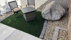 Une terrasse extérieure avec une table et deux chaises, entourée de gazon synthétique et d’un grand rocher.