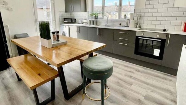 A modern kitchen with a wooden table and bench, grey cabinets, and a view through the window. - Minerva Close by Tŷ SA (Newport)