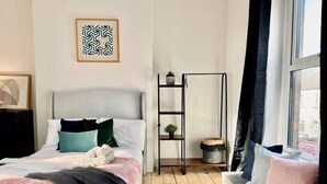 A modern bedroom with a bed, a bedside table, a window with curtains, a wall-mounted shelf with decorative items, and a framed picture. - Minerva Close by Tŷ SA (Newport)