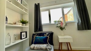 A modern bedroom with a bed, a white bedside table, a window with curtains, and a bookshelf with various items. - Minerva Close by Tŷ SA (Newport)