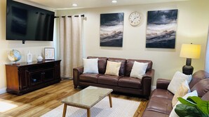 Una sala de estar con un sofá de cuero marrón, una mesa de centro de vidrio, un aparador de madera, un televisor de pantalla plana y dos relojes de pared.