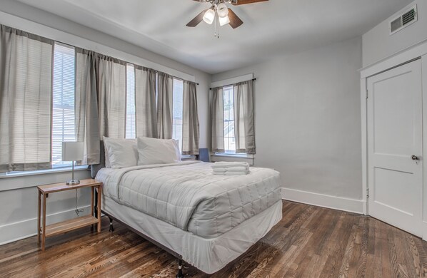 A bedroom with a bed, wooden floor, ceiling fan, and windows with curtains.