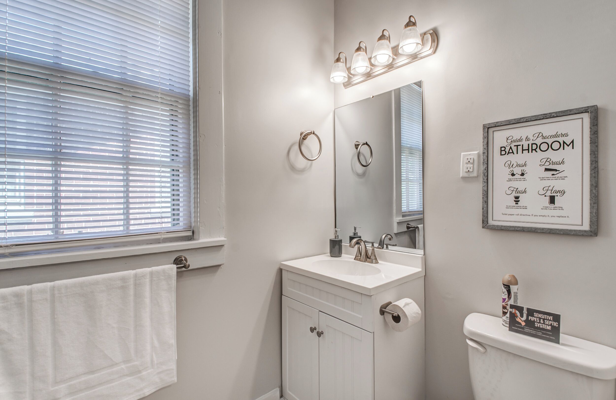 A bathroom with a sink, mirror, and toilet. There is a towel rack and a framed sign on the wall.