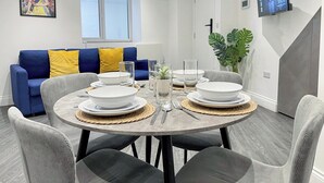 Un coin repas avec une table ronde dressée pour quatre personnes, un canapé bleu agrémenté de coussins jaunes, une grande fenêtre et un téléviseur fixé au mur.