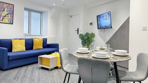 Un salon moderne avec un canapé bleu, une table à manger dressée pour quatre personnes et un téléviseur fixé au mur.