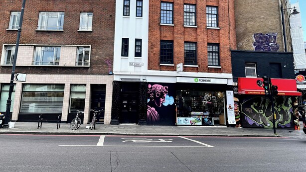 Une vue de rue présentant un mélange de bâtiments commerciaux, notamment un magasin appelé FIXHERE et un restaurant doté d’une terrasse.
