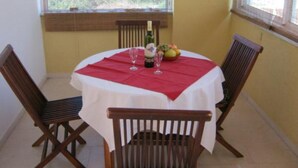 Un área de comedor con una mesa redonda cubierta por un mantel blanco y posavasos rojos, flanqueada por sillas de madera. Hay una botella y vasos sobre la mesa, y se ve un cuenco con fruta.