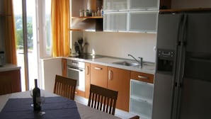 Cocina compacta con comedor, con una mesa dispuesta con una botella y fruta, un refrigerador y una ventana con cortinas.
