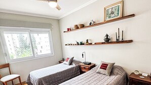 Une chambre avec deux lits, un plancher en bois, un ventilateur de plafond, une fenêtre avec vue et des étagères ornées d’objets décoratifs.