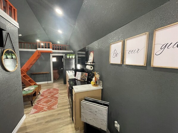 Et kompakt køkken med indbygget ovn, et lille spiseområde med bord og stole, og en trappe til en etage ovenover.