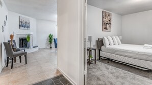 A modern, well-lit room with a bed, a small table with a lamp, and a chair. There is a fireplace and a plant in the background. - Near Parks Tranquil Fox Trails (Memphis)